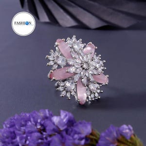 May include: A floral-shaped ring with pink and clear gemstones. The ring features a central cluster of clear stones surrounded by pink, petal-shaped stones. The ring is set on a silver-toned metal base. The word "FASHION" is in a white circle.