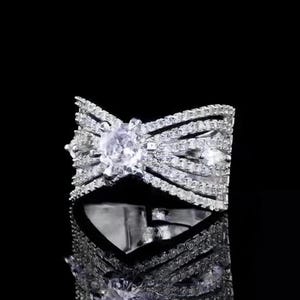 May include: A dazzling silver-toned ring with a bow-shaped design, encrusted with numerous sparkling clear gemstones. The ring features a large, central gemstone, and the design is reflected in the dark, glossy surface below.