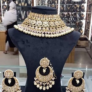 May include: A gold-toned necklace and earring set displayed on black velvet busts. The necklace is a choker style with multiple layers of gold filigree and clear stones, accented with small pearls. The earrings are crescent-shaped, also with gold filigree and pearls.