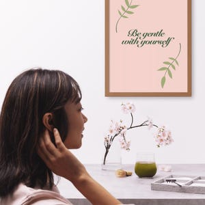 Op de afbeelding: Een ingelijste print met de tekst "Be gentle with yourself" in een scriptlettertype, tegen een roze achtergrond. Groene bladerillustraties staan ​​boven en onder. Een vrouw staat op de voorgrond.