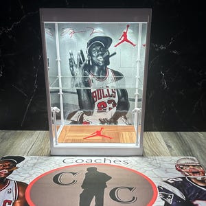 May include: A white display case featuring a graphic of Michael Jordan in a Chicago Bulls jersey. The case has a red Jumpman logo and a wooden floor design. The base has the text "Coaches Collectibles".