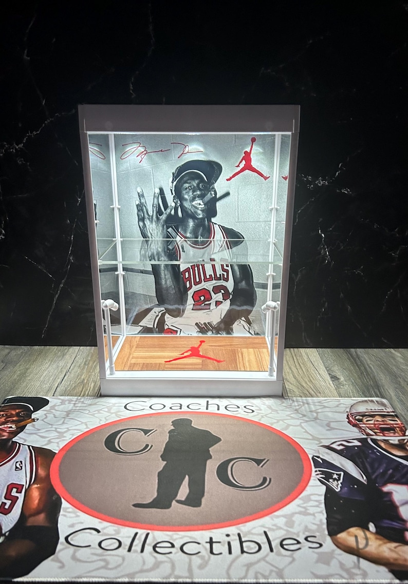 May include: A white display case featuring a graphic of Michael Jordan in a Chicago Bulls jersey. The case has a red Jumpman logo and a wooden floor design. The base has the text "Coaches Collectibles".