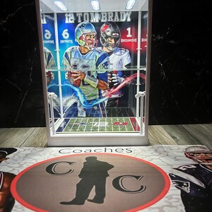 Puede incluir: Una vitrina blanca con un objeto de colecci&oacute;n de Tom Brady. La vitrina contiene una tarjeta de f&uacute;tbol con im&aacute;genes de Tom Brady con uniformes de los Patriots y los Buccaneers. La vitrina est&aacute; iluminada con luces superiores. La base de la vitrina tiene las palabras "Coaches Collectibles" y un logotipo.