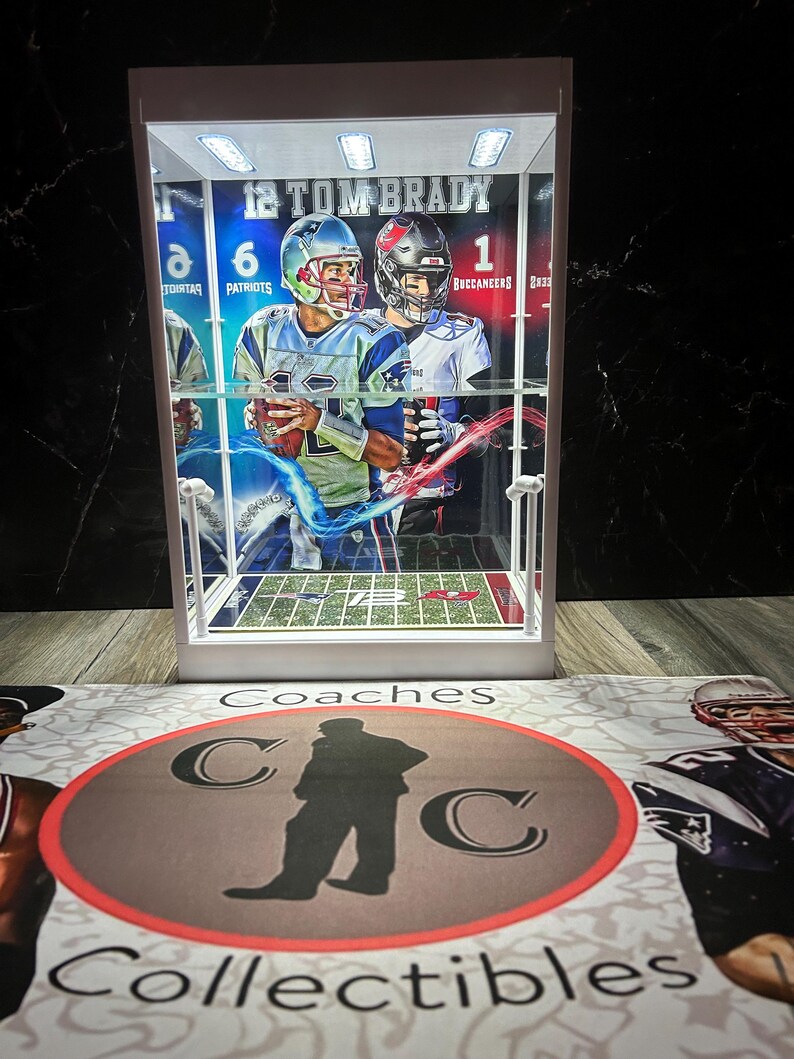 Puede incluir: Una vitrina blanca con un objeto de colecci&oacute;n de Tom Brady. La vitrina contiene una tarjeta de f&uacute;tbol con im&aacute;genes de Tom Brady con uniformes de los Patriots y los Buccaneers. La vitrina est&aacute; iluminada con luces superiores. La base de la vitrina tiene las palabras "Coaches Collectibles" y un logotipo.