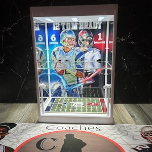 Puede incluir: Una vitrina blanca con una imagen de Tom Brady. La imagen muestra a Brady con un uniforme de los Patriots y de los Buccaneers. La vitrina tiene un campo de f&uacute;tbol americano en miniatura y el texto "12 TOM BRADY".