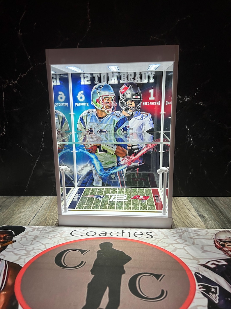 Puede incluir: Una vitrina blanca con una imagen de Tom Brady. La imagen muestra a Brady con un uniforme de los Patriots y de los Buccaneers. La vitrina tiene un campo de f&uacute;tbol americano en miniatura y el texto "12 TOM BRADY".