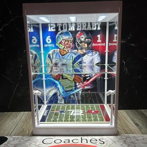 Puede incluir: Una vitrina blanca con un dise&ntilde;o de campo de f&uacute;tbol, que muestra im&aacute;genes de Tom Brady con uniformes de los Patriots y los Buccaneers. La vitrina tiene iluminaci&oacute;n interna y el texto "12 TOM BRADY" es visible.