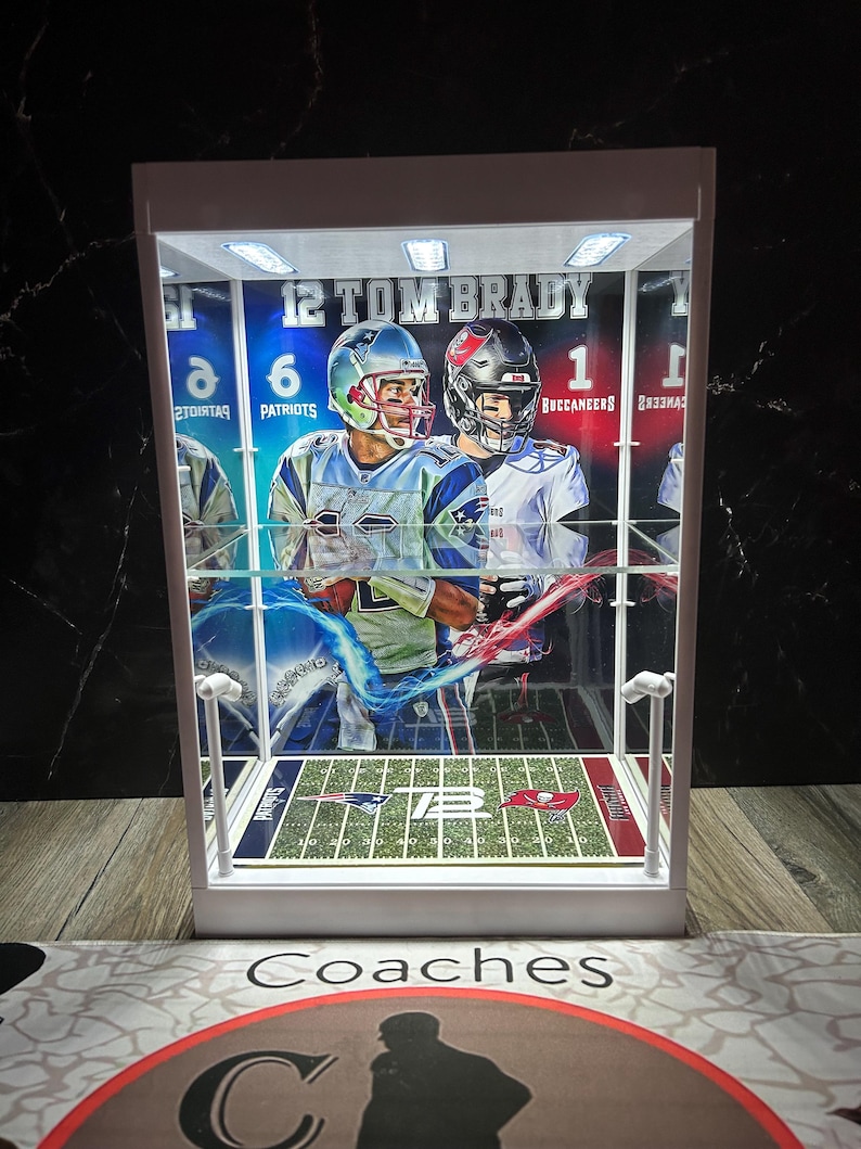 Puede incluir: Una vitrina blanca con un dise&ntilde;o de campo de f&uacute;tbol, que muestra im&aacute;genes de Tom Brady con uniformes de los Patriots y los Buccaneers. La vitrina tiene iluminaci&oacute;n interna y el texto "12 TOM BRADY" es visible.