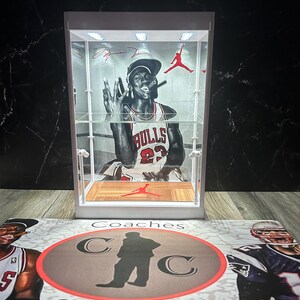 May include: A white display case with a black and white image of a basketball player in a Bulls jersey. The case has a red Jumpman logo and a wooden base. The case sits on a mat with the text "Coaches Collectibles".