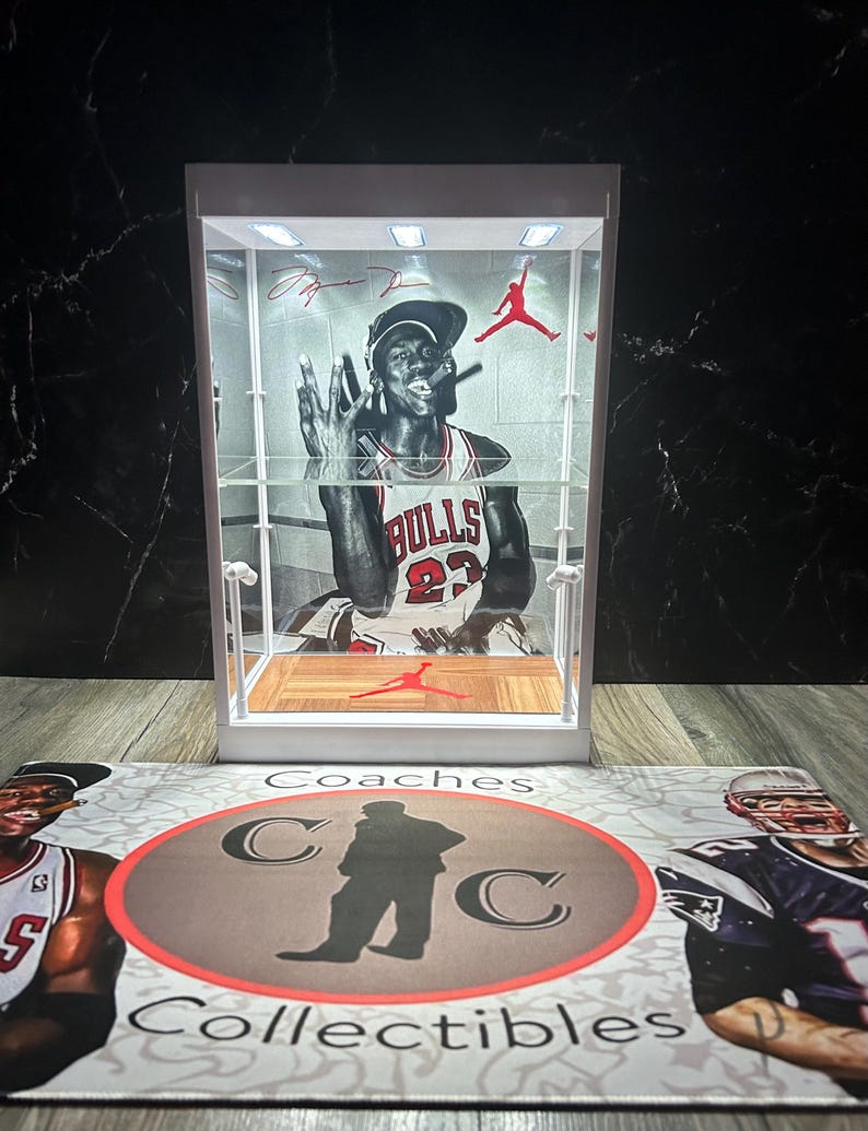 May include: A white display case with a black and white image of a basketball player in a Bulls jersey. The case has a red Jumpman logo and a wooden base. The case sits on a mat with the text "Coaches Collectibles".