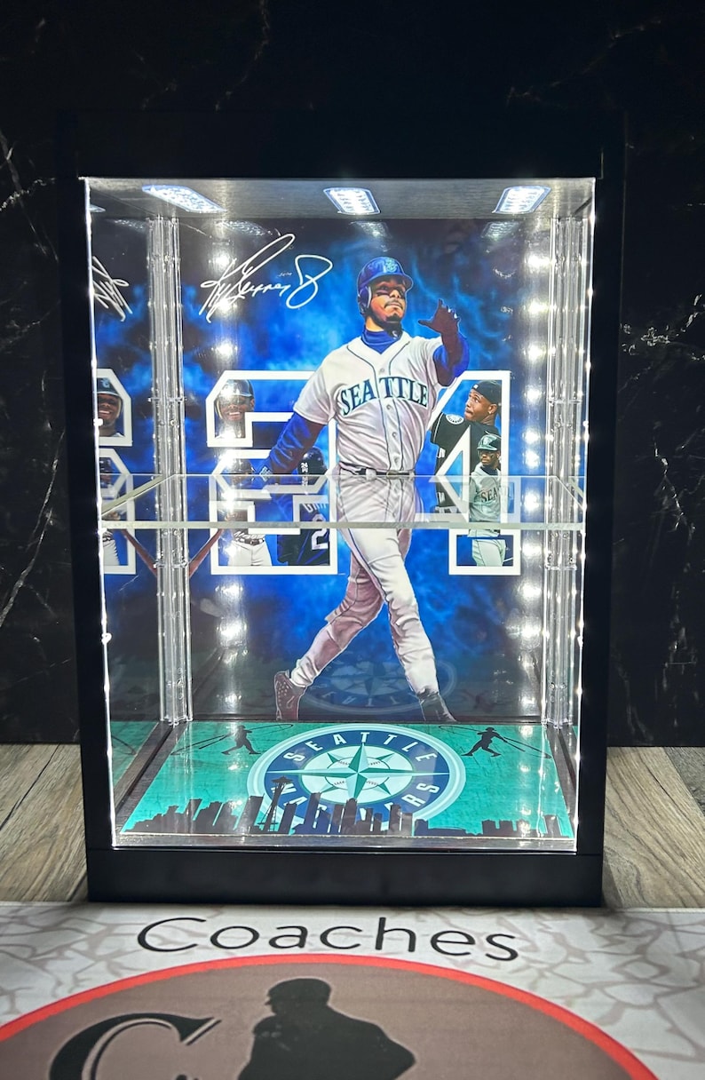 May include: A lit display case featuring a baseball player in a Seattle Mariners uniform. The case has a black frame and a green and blue baseball field design at the base. The player's image is surrounded by a blue and white background.