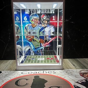 Puede incluir: Una vitrina blanca con un dise&ntilde;o de campo de f&uacute;tbol y im&aacute;genes de Tom Brady con uniformes de los Patriots y los Buccaneers. La vitrina tiene luces integradas y el texto "12 TOM BRADY" es visible. La base tiene la palabra "Coaches" y un logotipo circular.