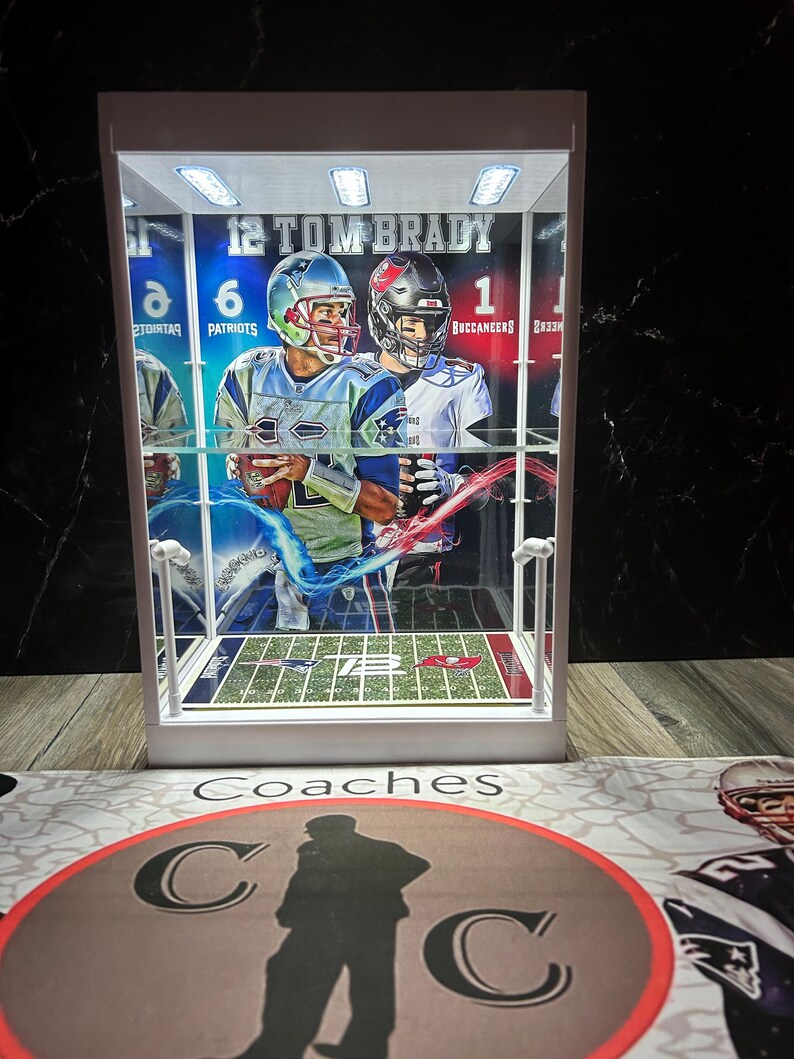 Puede incluir: Una vitrina blanca con un dise&ntilde;o de campo de f&uacute;tbol y im&aacute;genes de Tom Brady con uniformes de los Patriots y los Buccaneers. La vitrina tiene luces integradas y el texto "12 TOM BRADY" es visible. La base tiene la palabra "Coaches" y un logotipo circular.