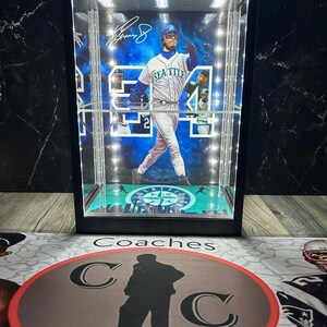 May include: A black display case featuring a baseball player in a Seattle Mariners uniform. The case has integrated lighting and a base with a cityscape design. The player's signature is visible. The case rests on a mat with the word "Coaches" and a silhouette.