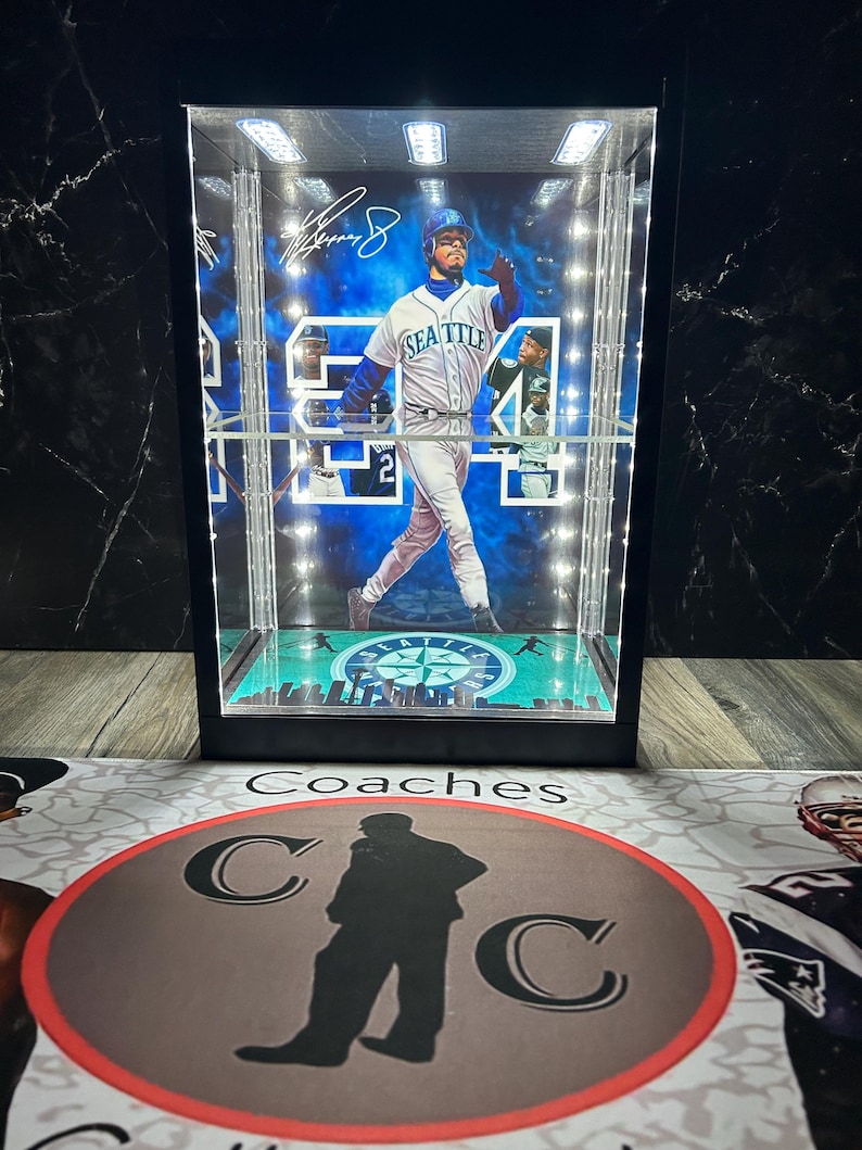 May include: A black display case featuring a baseball player in a Seattle Mariners uniform. The case has integrated lighting and a base with a cityscape design. The player's signature is visible. The case rests on a mat with the word "Coaches" and a silhouette.