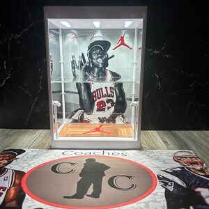 May include: A white display case featuring a black and white image of Michael Jordan in a Chicago Bulls jersey. The case has interior lighting and a red Jumpman logo. The base has a mat with the text "Coaches Collectibles" and sports figures.