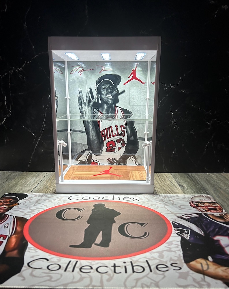 May include: A white display case featuring a black and white image of Michael Jordan in a Chicago Bulls jersey. The case has interior lighting and a red Jumpman logo. The base has a mat with the text "Coaches Collectibles" and sports figures.