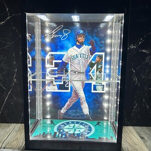 May include: A display case featuring a baseball player in a Seattle Mariners uniform. The case has a black frame, a clear front, and is illuminated with LED lights. The player's signature is visible. The base of the case has a green and blue design.