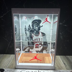 May include: A white display case with a black and white image of Michael Jordan in his Chicago Bulls jersey. The case has a wooden floor design with a red Jumpman logo and red accents. The case has a clear front and shelves.