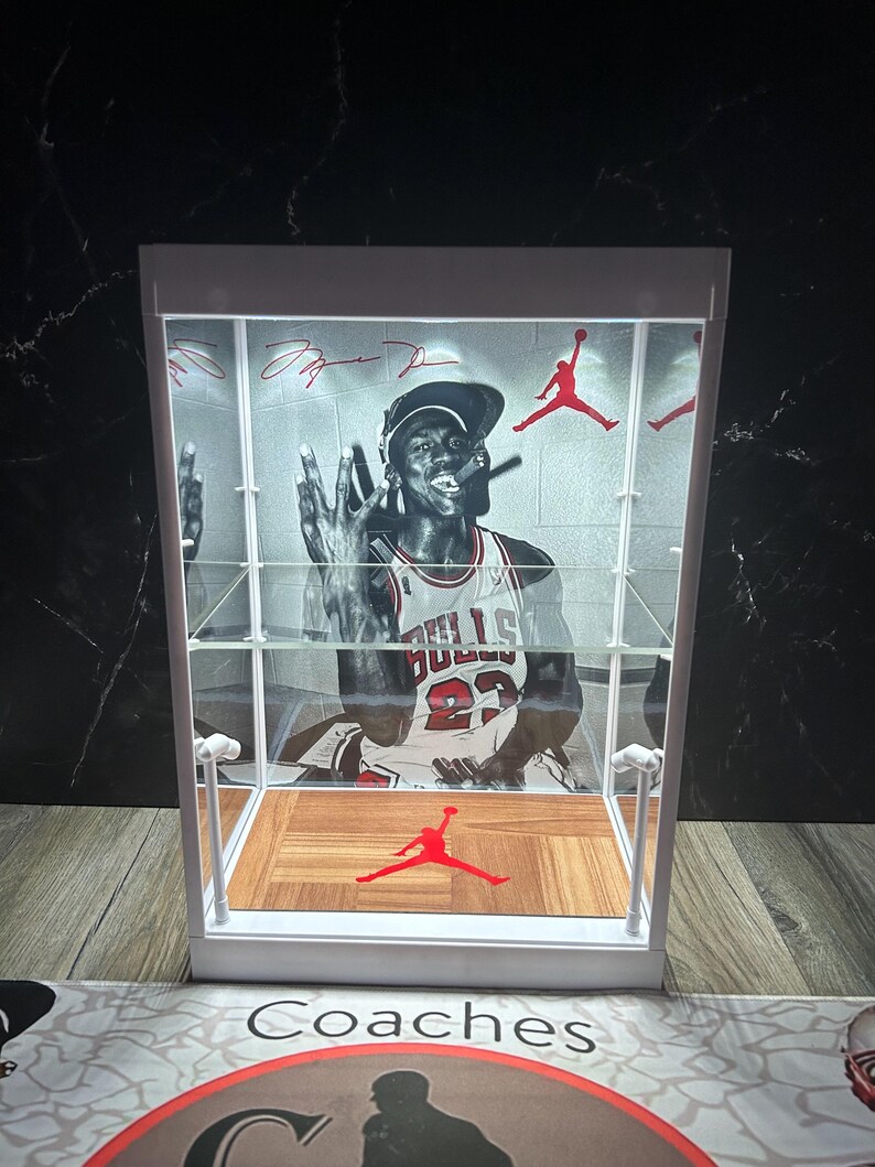 May include: A white display case with a black and white image of Michael Jordan in his Chicago Bulls jersey. The case has a wooden floor design with a red Jumpman logo and red accents. The case has a clear front and shelves.