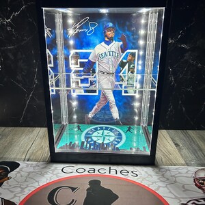 May include: A black display case featuring a baseball player in a Seattle Mariners uniform, number 54. The case has internal lighting and a green and blue baseball field design at the base. The case is a collectible item.