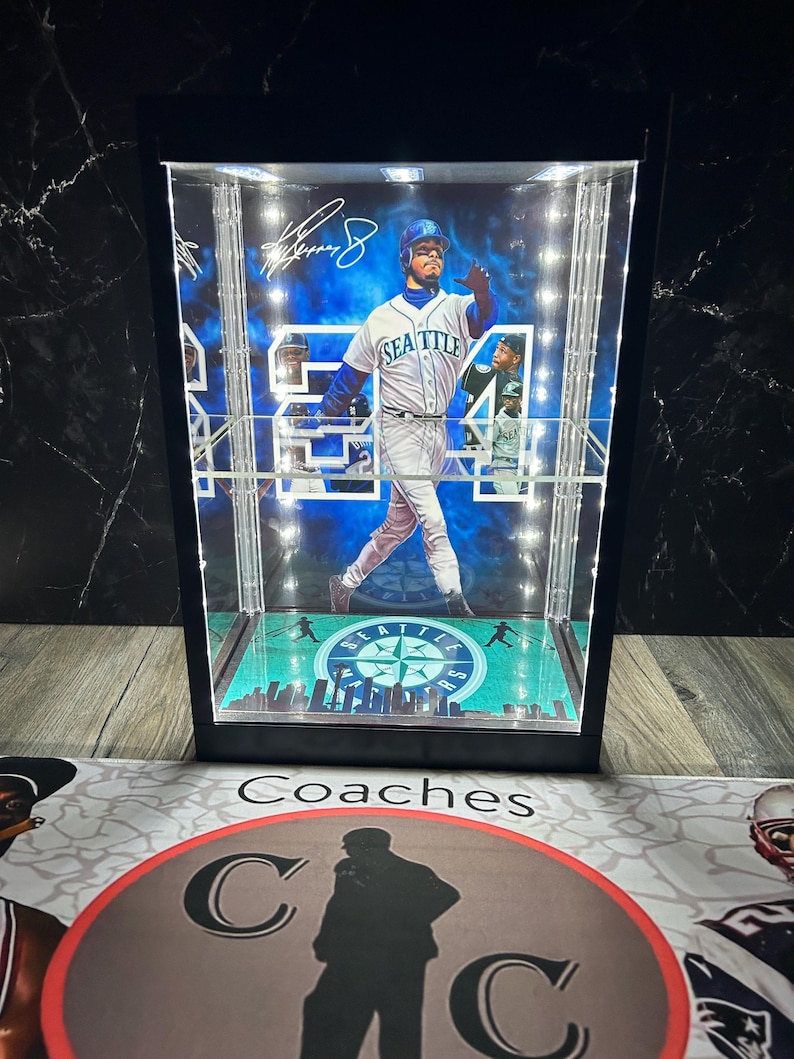 May include: A black display case featuring a baseball player in a Seattle Mariners uniform, number 54. The case has internal lighting and a green and blue baseball field design at the base. The case is a collectible item.