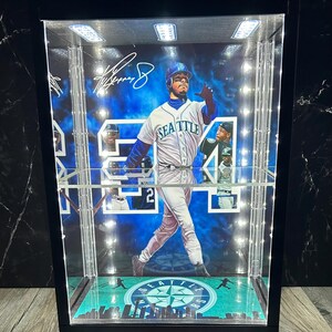 May include: A black display case featuring a baseball player in a Seattle Mariners uniform. The case has built-in lights and a green and blue base with the team logo. The player's signature is visible. The case is designed to showcase memorabilia.