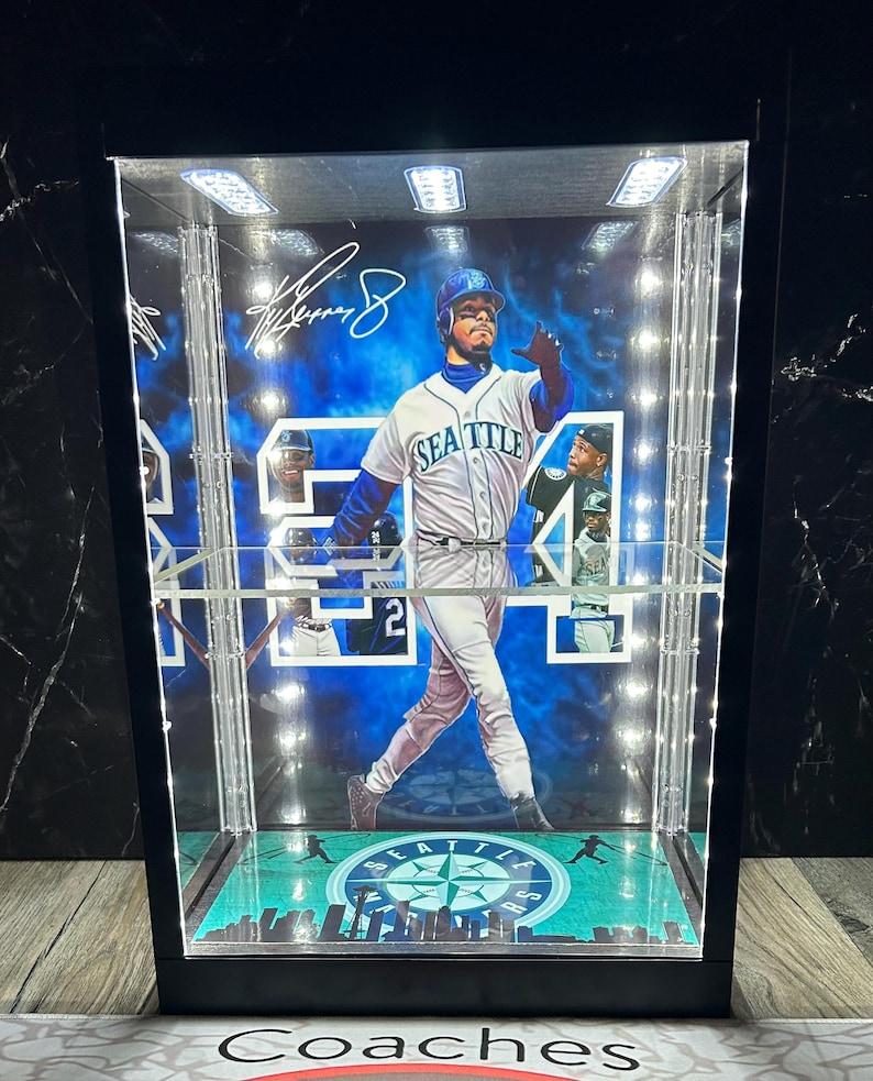 May include: A black display case featuring a baseball player in a Seattle Mariners uniform. The case has built-in lights and a green and blue base with the team logo. The player's signature is visible. The case is designed to showcase memorabilia.