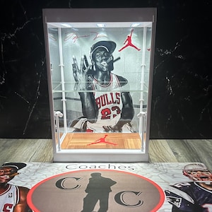 May include: A white display case featuring a black and white image of Michael Jordan in a Chicago Bulls jersey. The case has a wooden floor with a red Jumpman logo. Red accents and a Jumpman logo are visible.