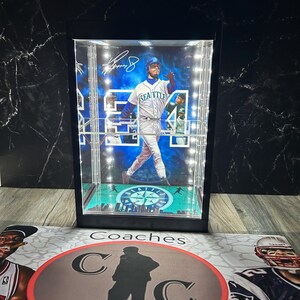 May include: A baseball player figurine in a lit display case. The player wears a white and blue uniform with "Seattle" on the front. The case has a black frame and a green base with a team logo. The case is on a surface with the word "Coaches".
