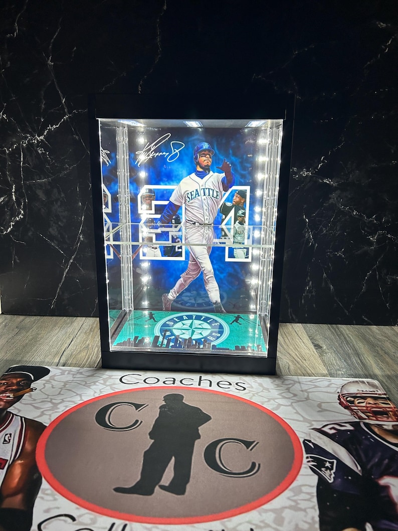 May include: A baseball player figurine in a lit display case. The player wears a white and blue uniform with "Seattle" on the front. The case has a black frame and a green base with a team logo. The case is on a surface with the word "Coaches".