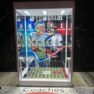 Puede incluir: Una vitrina blanca con una tarjeta de f&uacute;tbol de Tom Brady. La tarjeta muestra a Brady con un uniforme de los Patriots y de los Buccaneers. La vitrina tiene un dise&ntilde;o de campo de f&uacute;tbol en miniatura y est&aacute; iluminada con peque&ntilde;as luces.
