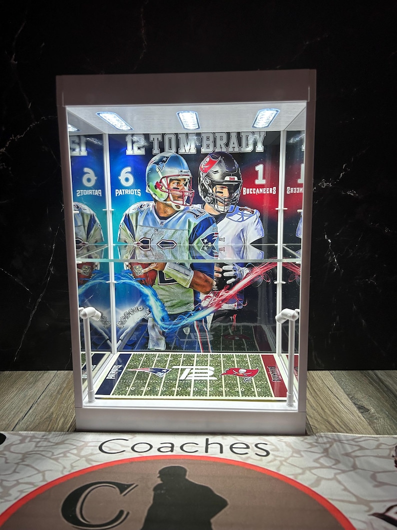 Puede incluir: Una vitrina blanca con una tarjeta de f&uacute;tbol de Tom Brady. La tarjeta muestra a Brady con un uniforme de los Patriots y de los Buccaneers. La vitrina tiene un dise&ntilde;o de campo de f&uacute;tbol en miniatura y est&aacute; iluminada con peque&ntilde;as luces.