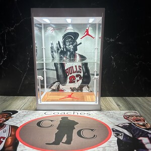 May include: A white display case featuring a black and white image of a basketball player. The case has a wooden floor and a red logo. The background is a black marble-effect surface. The base has the words "Coaches Collectibles".