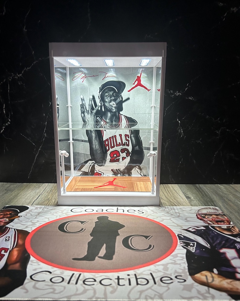 May include: A white display case featuring a black and white image of a basketball player. The case has a wooden floor and a red logo. The background is a black marble-effect surface. The base has the words "Coaches Collectibles".