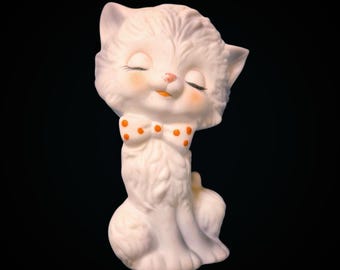 May include: A white ceramic cat figurine with a textured, fur-like surface. The cat has closed eyes, pink-tinted cheeks, and a small orange nose. It wears a white bow tie featuring orange polka dots around its neck, with a seated posture.