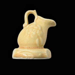 May include: A small, cream-colored ceramic creamer with a handle and spout. The creamer has a decorative floral pattern on the side and sits on a rectangular base. The background is black.