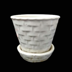 May include: A white ceramic planter with a textured basket weave design. The planter has a slightly flared shape and sits on a matching saucer. The background is solid black.