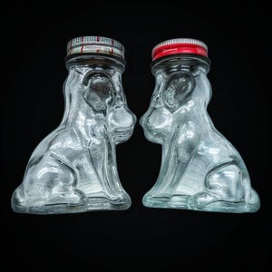 May include: Two clear glass dog-shaped bottles with metal lids. One lid is red and the other is silver. The bottles are side-by-side against a black background. The bottles are likely vintage or collectible items.