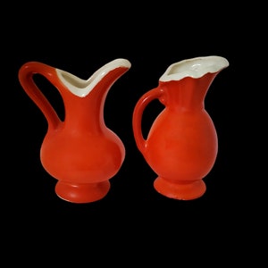 May include: Two small orange ceramic pitchers with white rims. The pitchers have a classic design and are perfect for a farmhouse or country kitchen.