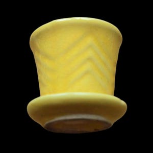 May include: A yellow ceramic top hat-shaped container with a wide brim and a slightly textured surface. The hat has a chevron pattern on the sides and a dark brown interior. The background is black.