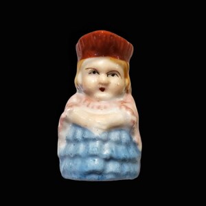 May include: A small ceramic figurine of a girl wearing a red hat and a blue dress. The figurine has a painted face with a serious expression.