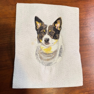 May include: A rectangular piece of light gray felt fabric featuring a detailed machine embroidery of a dog. The dog has black, white, and tan fur, pointed ears, and a focused expression. The embroidery uses varied thread colors to create texture.