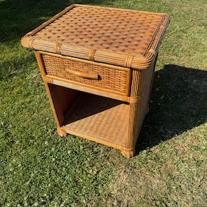 May include: A light brown wicker nightstand featuring a square top with a woven pattern, a single drawer with a curved handle, and an open storage shelf at the base. The piece is constructed from natural rattan material with a consistent weave.