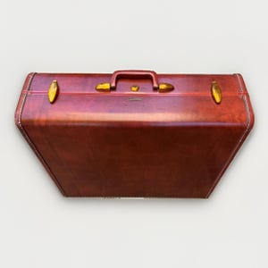 May include: A rectangular, deep reddish-brown suitcase with a smooth, leather-like finish. It features a central top handle, two gold-toned metal latches on either side, and a central locking mechanism. The edges are reinforced with white stitching.