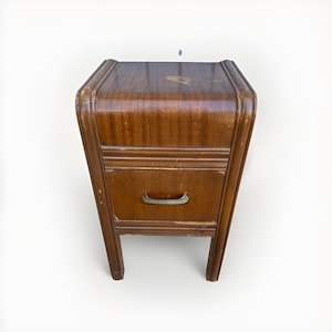 May include: A brown wooden nightstand featuring a curved top edge, a single drawer with a metallic handle, and four straight legs. The wood grain is visible across the surface, which shows signs of wear and minor surface scratches throughout.