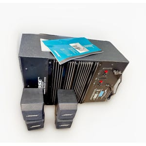 May include: A black Bose Powered Speaker System with two satellite speakers. The system includes a subwoofer with a ribbed front panel and a blue and white manual. The Bose logo is visible on the speakers.