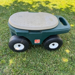 May include: A dark green plastic rolling garden seat with a beige, perforated, oval-shaped lid. The base features four black wheels with white centers and a handle integrated into the side. The brand name Step2 is printed in red on the side.