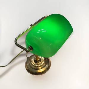 May include: A banker's lamp featuring a vibrant green glass shade and a polished brass base. The lamp has a curved metal arm that supports the cylindrical shade, which is attached to a circular, fluted brass stand with a power cord attached.