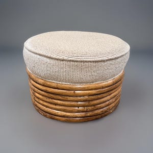 May include: A round ottoman with a light beige upholstered top and a natural rattan base. The base is constructed of horizontal, curved wooden slats. The ottoman is a neutral color and would complement many decor styles.